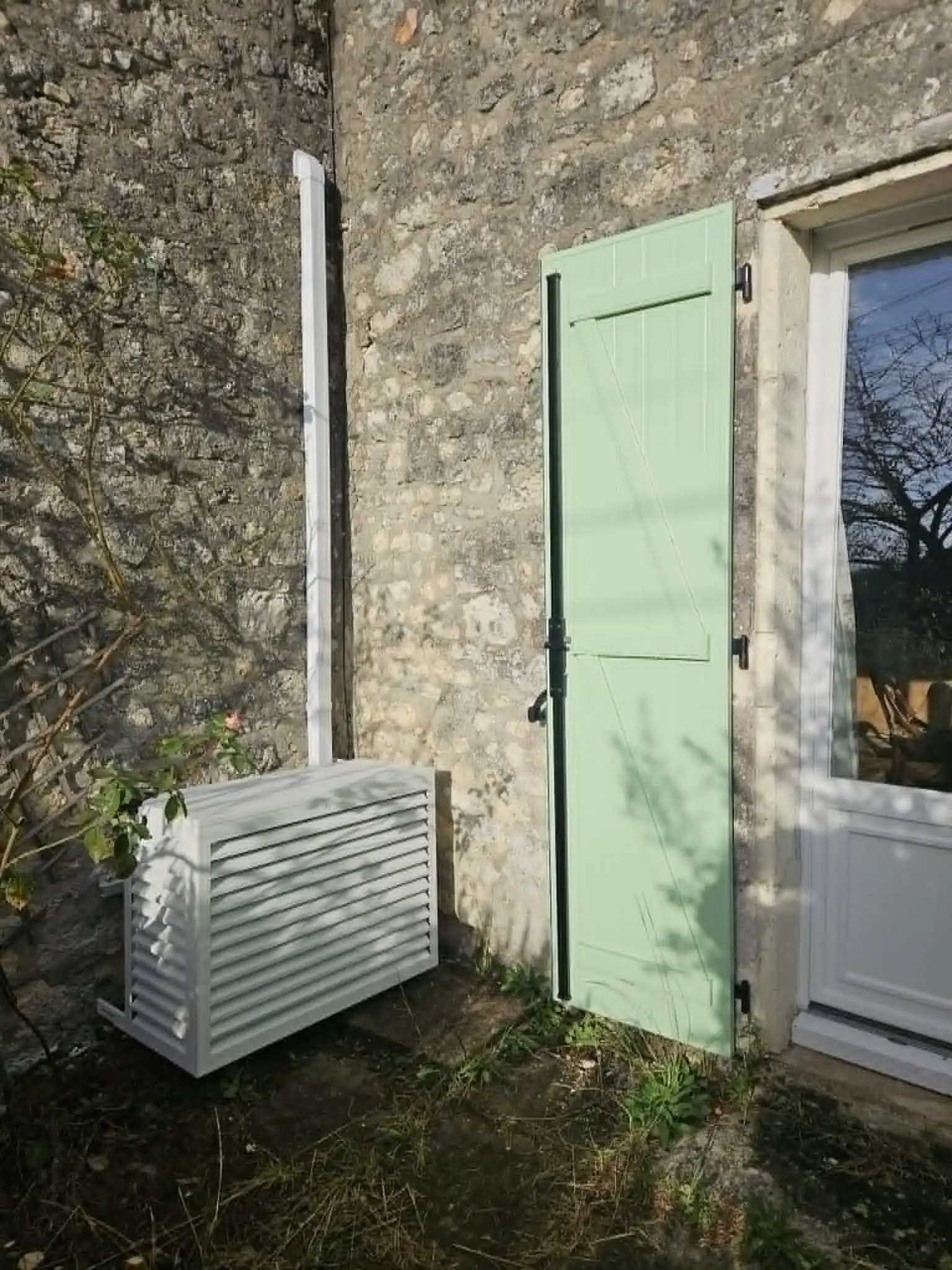 Unité extérieure cachée derrière un coffre blanc, maison en pierre avec volet vert